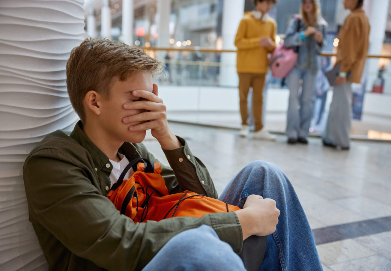 Friendship problem and bad situation at shopping mall. Unhappy teenage boy feeling lonely, unaccepted, rejected by friends. Young boy sitting with covered face with hand